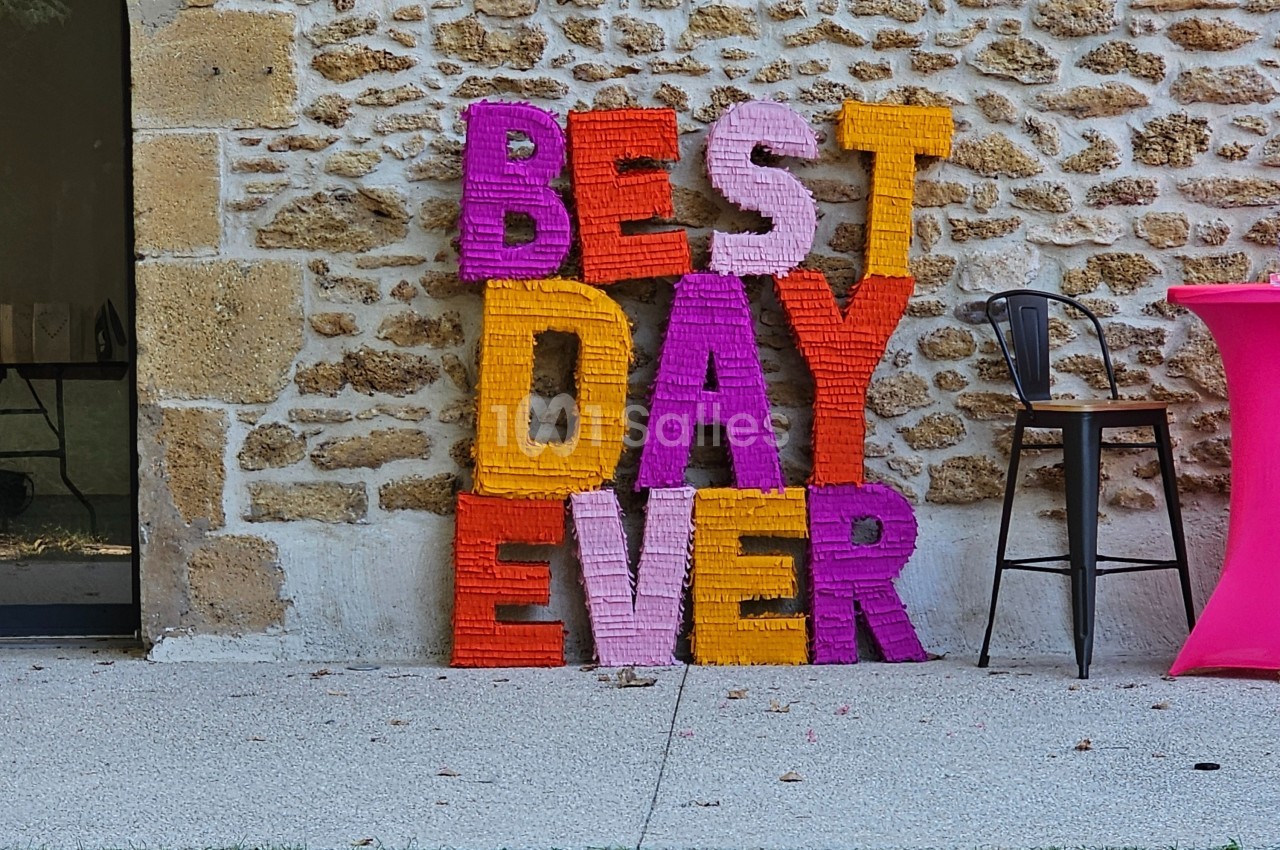 Lettres colorées formant ’Best Day Ever’ appuyées contre un mur en pierre, près d'une table et d'une chaise.