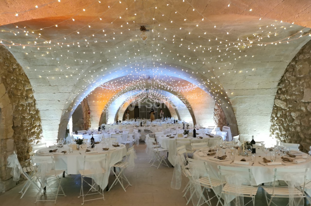 Salle voûtée en pierre décorée de guirlandes lumineuses, tables dressées avec nappes blanches pour un événement.