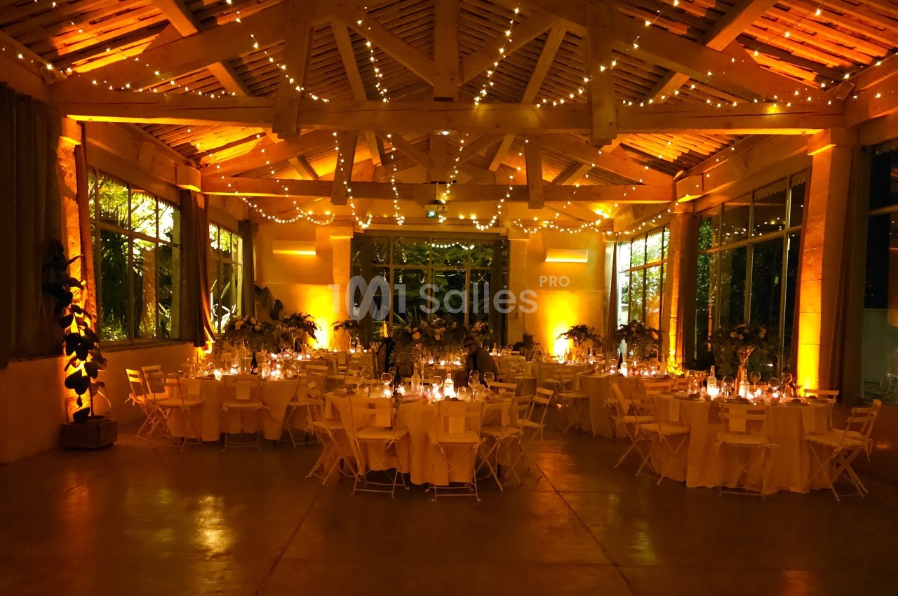 Salle de réception illuminée par des guirlandes lumineuses, avec des tables rondes décorées pour un événement festif.