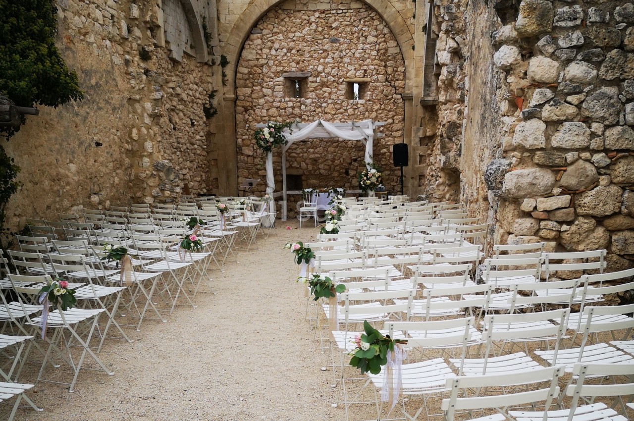 Chaises blanches alignées dans une cour en pierre, décorée pour une cérémonie avec un autel fleuri sous une arche blanche.