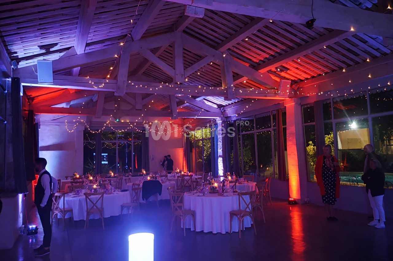 Salle de réception décorée avec des guirlandes lumineuses, tables rondes dressées et ambiance tamisée.