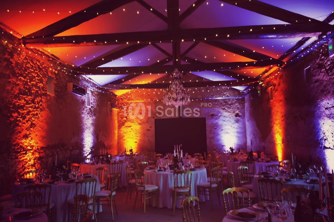 Salle de réception en pierre éclairée par des lumières colorées, avec tables rondes dressées pour un événement.