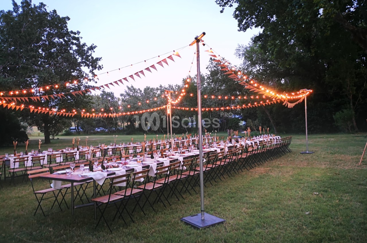 Tables longues dressées en extérieur avec guirlandes lumineuses et fanions suspendus, entourées d'arbres.