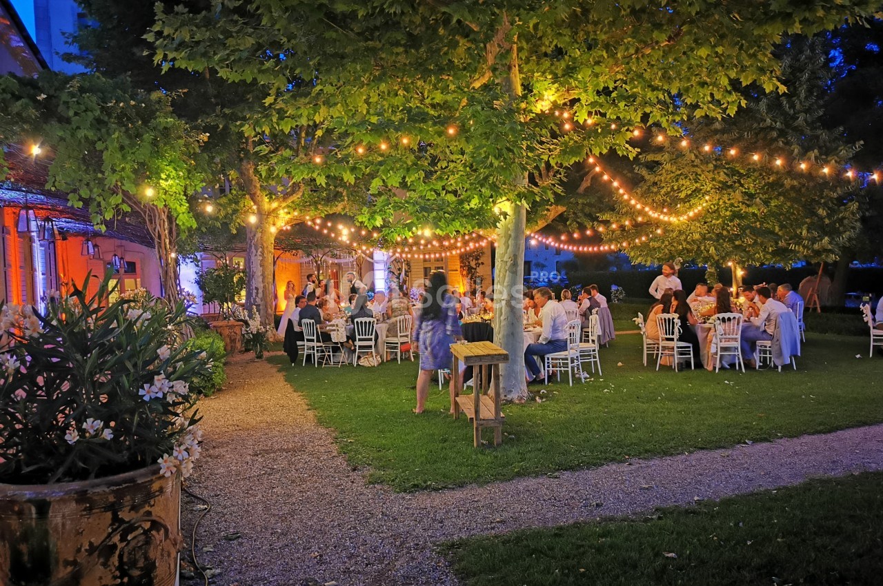 Dîner en plein air sous des guirlandes lumineuses, avec des invités assis à des tables dans un jardin en soirée.