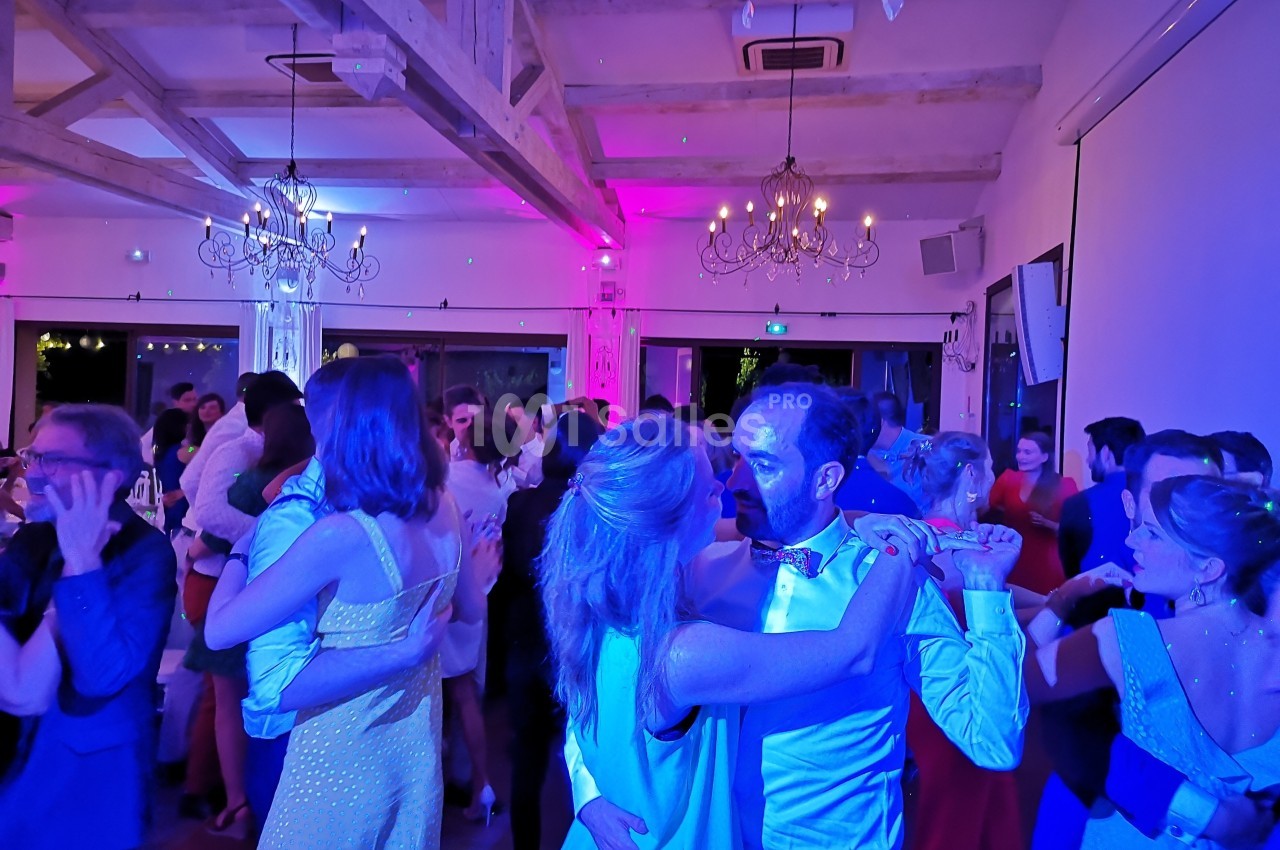 Des couples dansent dans une salle éclairée par des lumières colorées, avec des poutres apparentes et des lustres suspendus.
