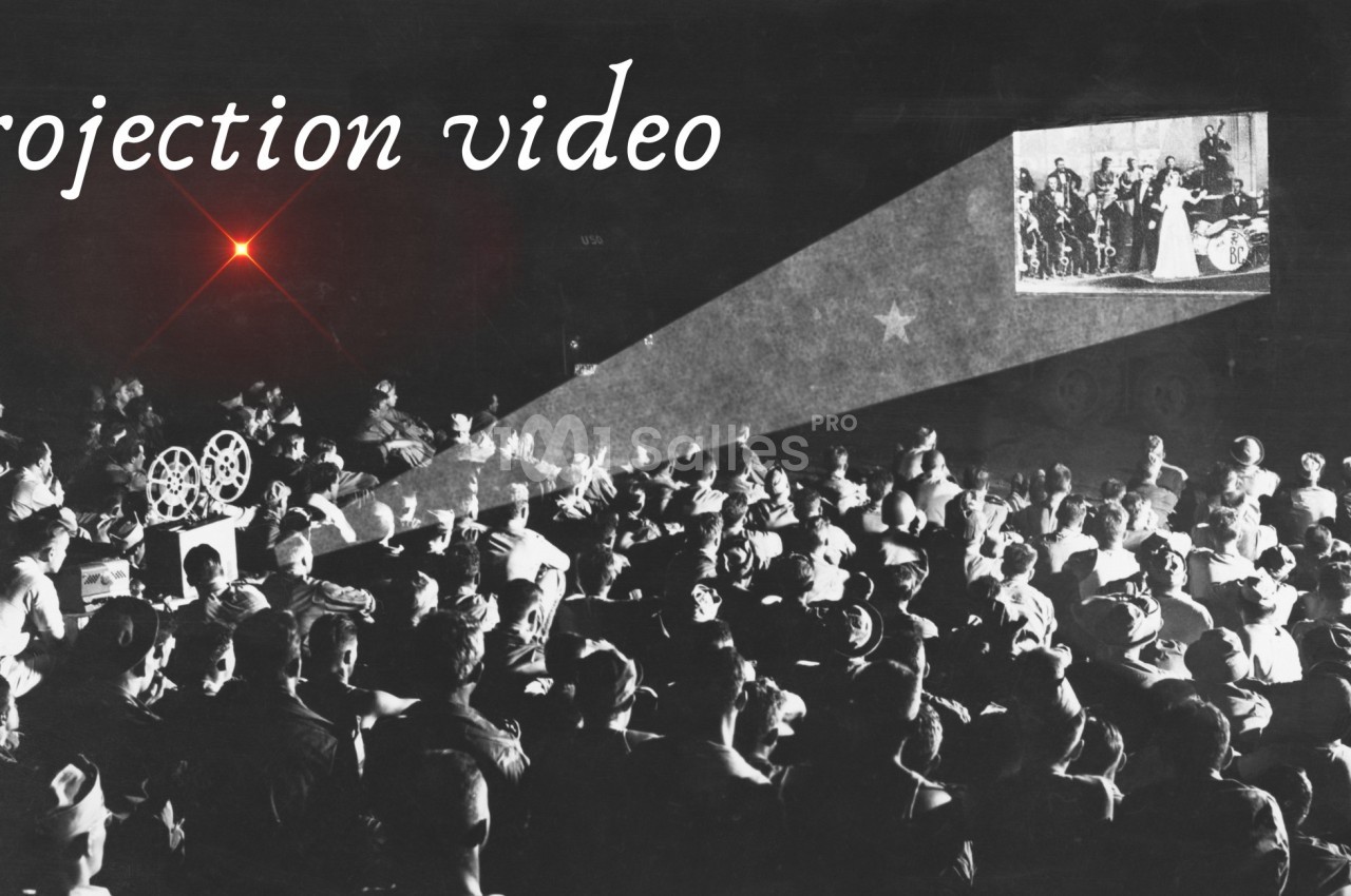 Projection d'un film en noir et blanc devant un public nombreux, avec un projecteur visible à gauche.