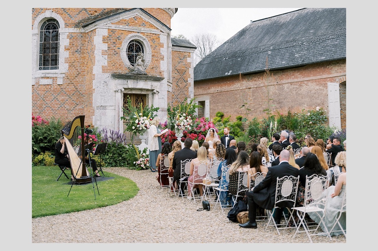 Cérémonie de mariage en plein air devant une chapelle en briques, avec invités assis et une harpiste jouant.