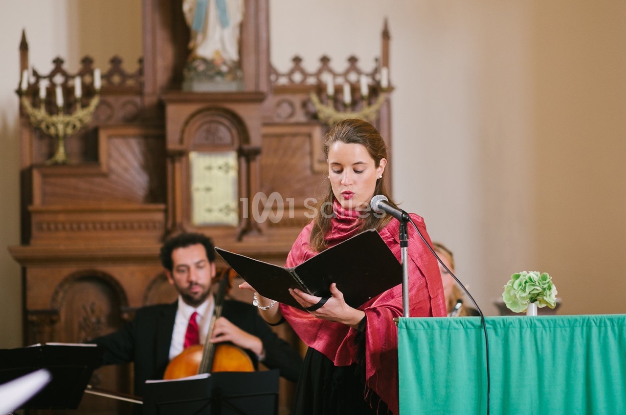 Une femme chante devant un micro en lisant une partition, accompagnée de musiciens dans un décor d'église.