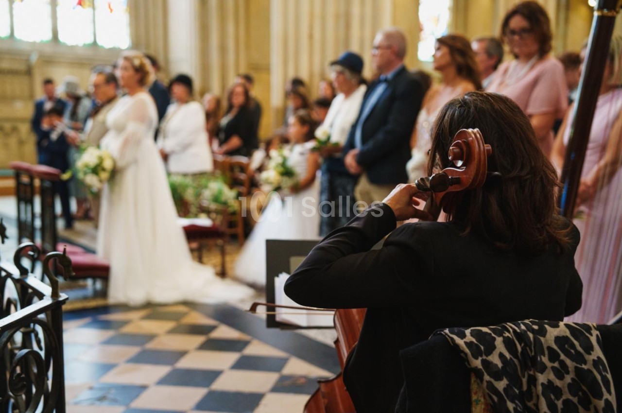 Une violoncelliste joue lors d'une cérémonie dans une église, entourée d'invités debout et de mariés à l'arrière-plan.
