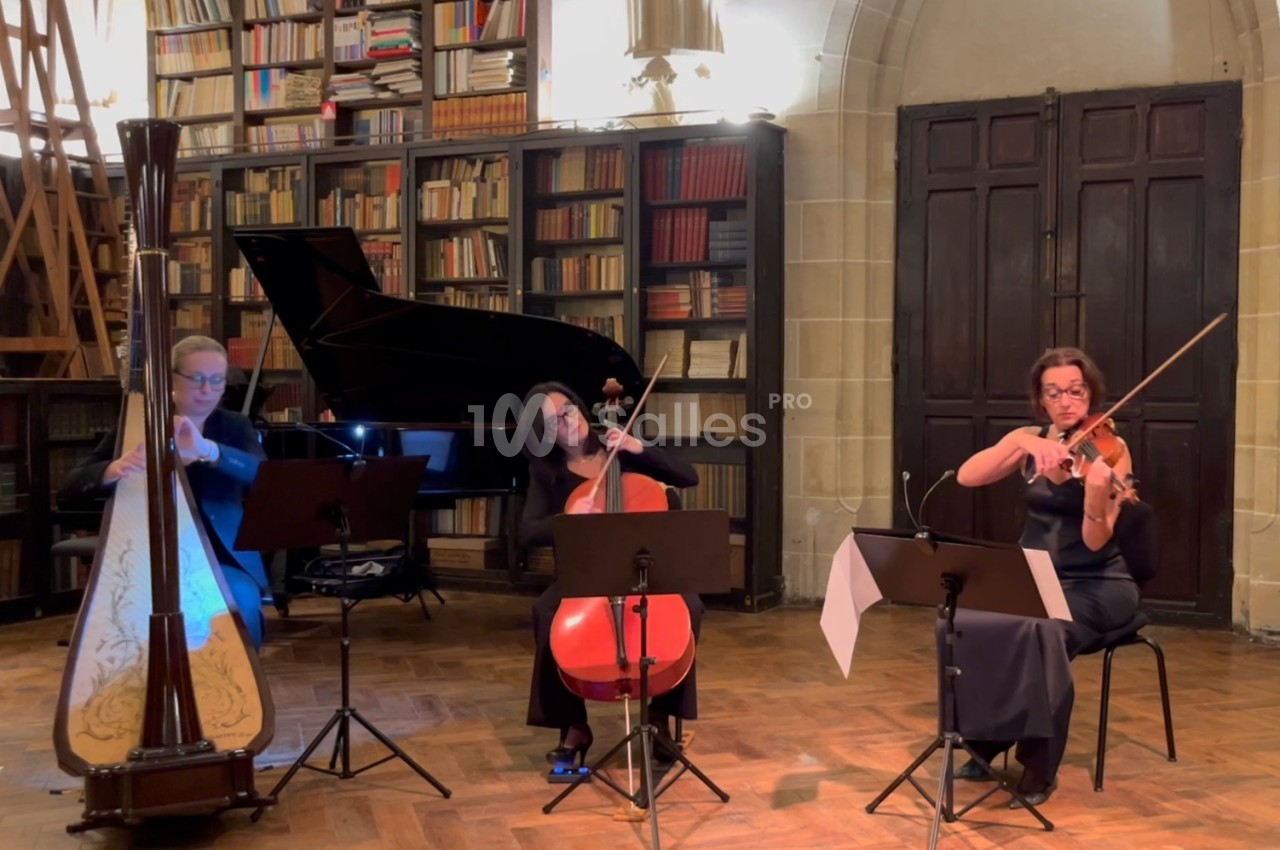 Trois musiciennes jouent de la harpe, du violoncelle et du violon dans une salle avec des étagères remplies de livres.
