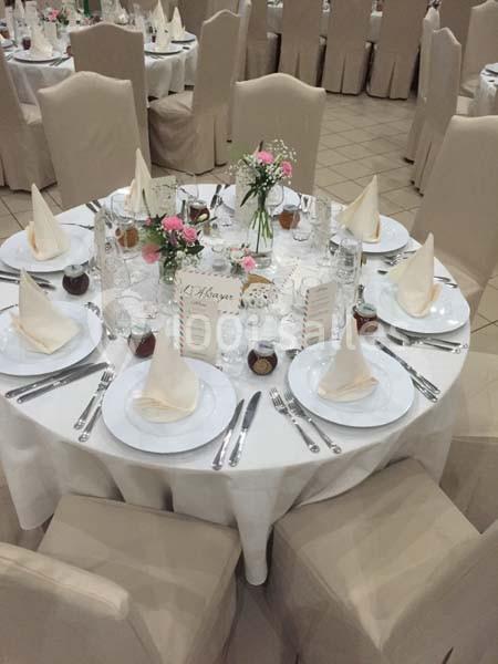 Table ronde dressée pour un événement, avec nappes blanches, serviettes pliées, couverts, verres et décorations florales.