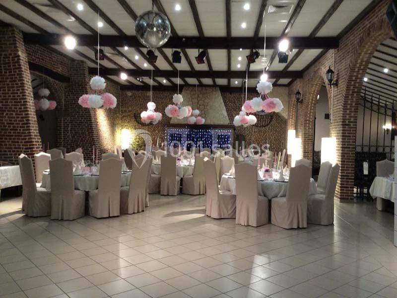 Salle de réception décorée avec des tables rondes, nappes blanches, chaises houssées et guirlandes de pompons suspendus.