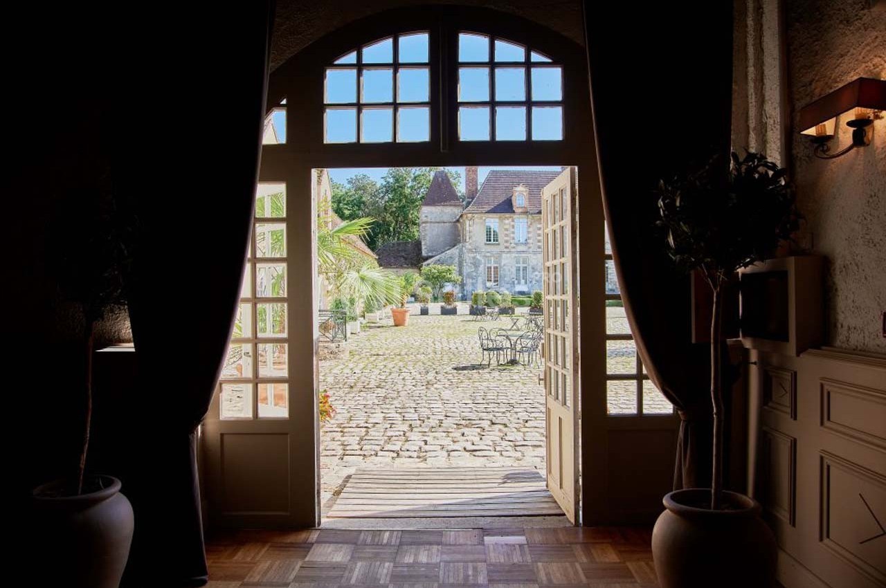 Vue depuis l'intérieur d'une porte ouverte sur une cour pavée ensoleillée avec des tables et un bâtiment ancien en arrière…
