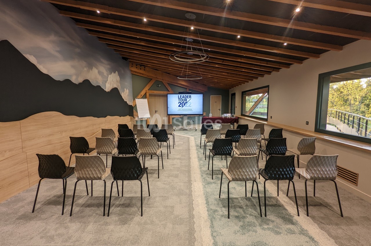 Salle de conférence lumineuse avec des rangées de chaises vides, un écran de présentation et un mur décoré de montagnes.