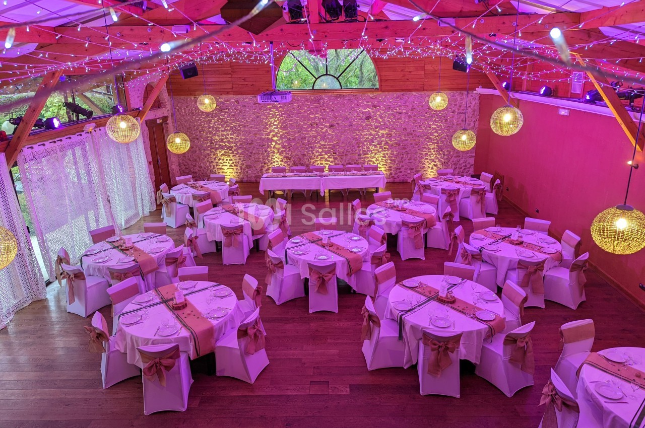 Salle de réception décorée pour un mariage, avec tables rondes dressées, chaises ornées et éclairage chaleureux.