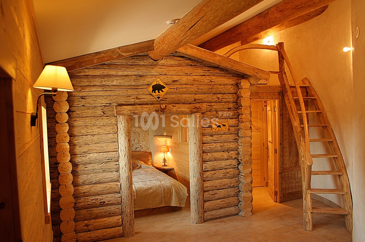 Chambre rustique avec murs en rondins, lit éclairé par une lampe et mezzanine accessible par une échelle en bois.