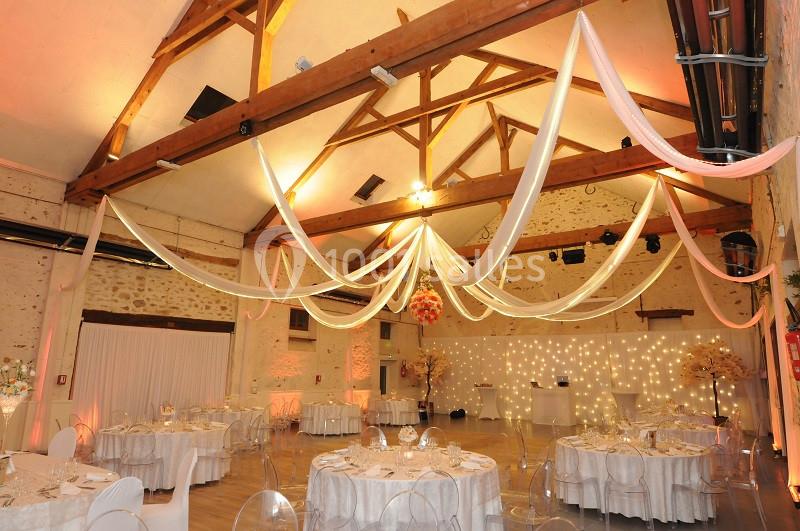 Salle de réception décorée avec des tables rondes, nappes blanches et drapés suspendus aux poutres en bois apparentes.