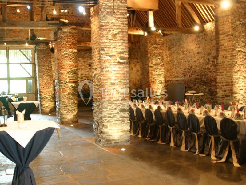 Salle en pierre avec poutres apparentes, tables dressées pour un événement et lumière naturelle entrant par une grande baie…