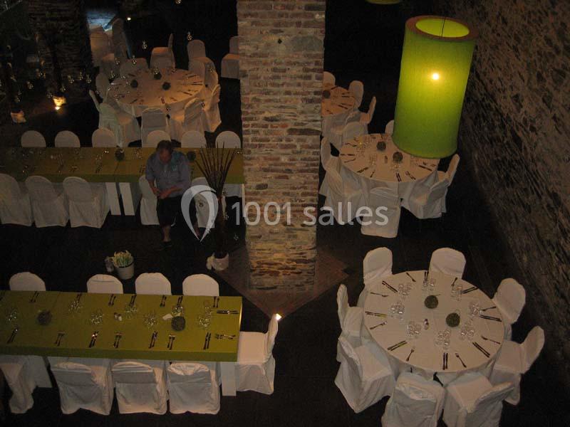 Salle de réception avec des tables rondes et rectangulaires dressées, chaises blanches et grandes lampes vertes.