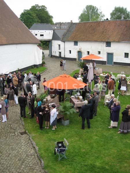 Groupe de personnes rassemblées dans une cour pavée entourée de bâtiments anciens, avec des parasols orange.