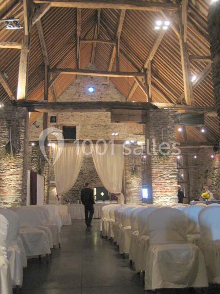 Salle en pierre avec poutres apparentes, rangées de chaises blanches et rideaux clairs au fond.