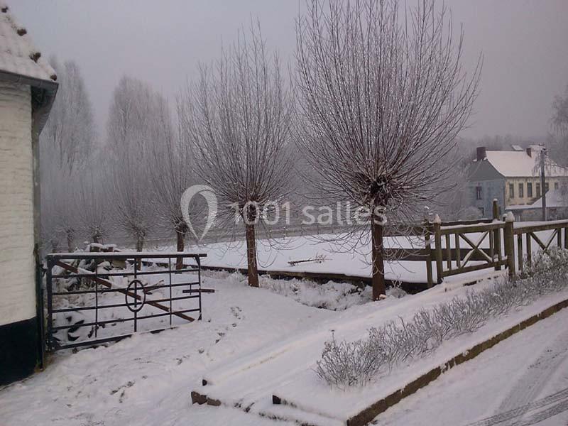 Paysage hivernal avec arbres dénudés, sol enneigé, clôture en bois et maisons en arrière-plan sous un ciel gris.