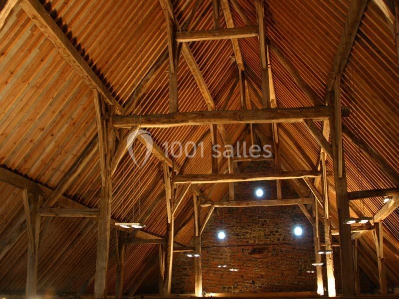 Charpente en bois apparente d'un grenier ou d'une grange, avec un toit en tuiles et un mur en briques au fond.