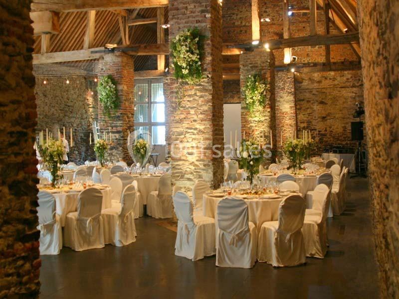Salle de réception en pierre avec tables rondes décorées, chaises blanches et arrangements floraux suspendus.