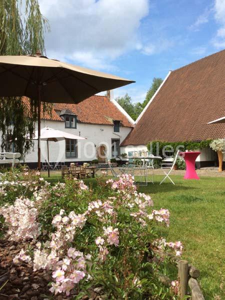 Jardin avec fleurs roses, tables et chaises sous des parasols, devant des bâtiments aux toits en tuiles rouges.