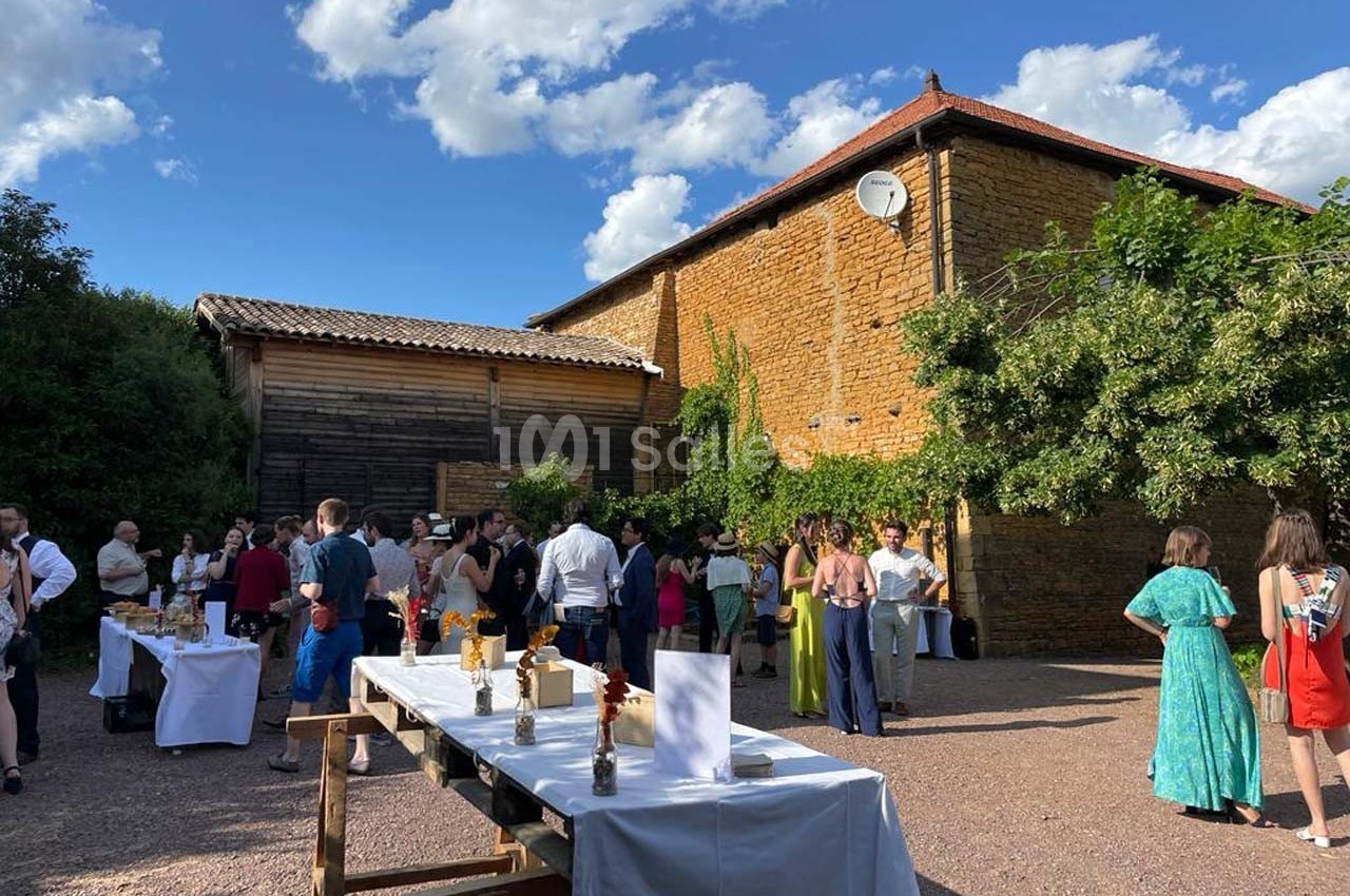 Groupe de personnes rassemblées dans une cour ensoleillée avec tables dressées, près de bâtiments en pierre et bois.