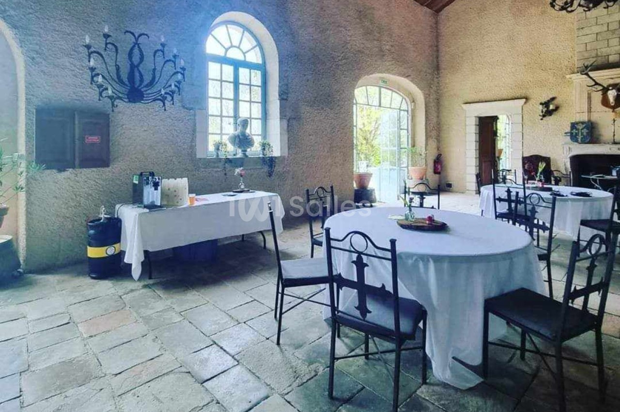 Salle en pierre avec tables rondes dressées, nappes blanches, buffet à boissons et grande fenêtre laissant entrer la lumière.