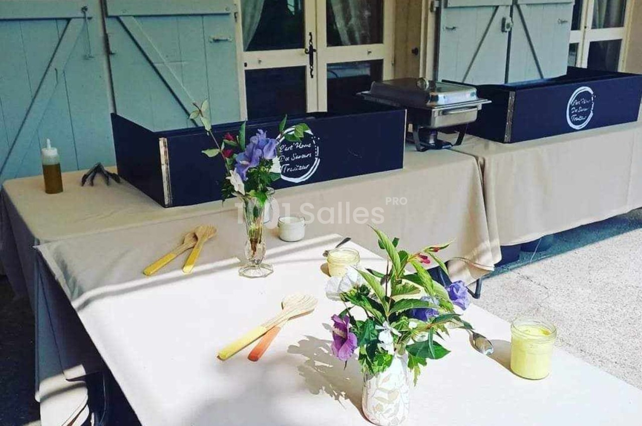 Tables dressées avec nappes beiges, décorées de bouquets de fleurs et d'ustensiles en bois, devant des volets bleus.
