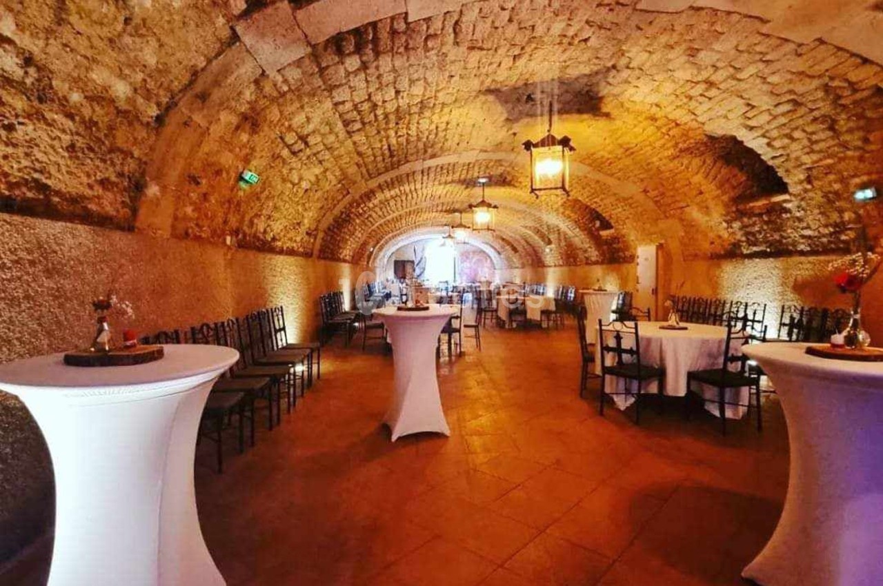 Salle voûtée en pierre avec tables rondes dressées et mange-debout, éclairée par des lanternes suspendues.