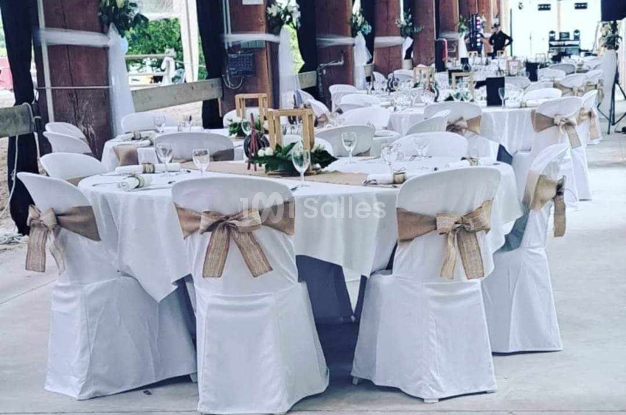 Salle décorée pour un événement avec des tables rondes dressées, nappes blanches et chaises ornées de nœuds en toile de jute.
