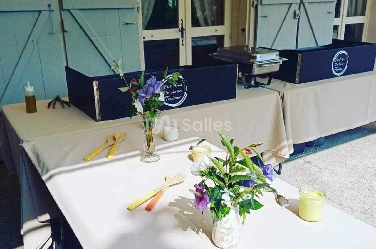 Tables dressées avec nappes beiges, décorées de bouquets de fleurs et d'ustensiles en bois, devant des volets bleus.