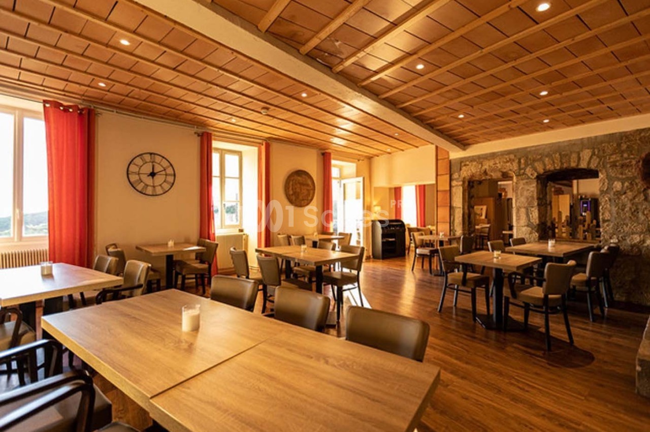 Salle de restaurant lumineuse avec tables en bois, chaises modernes, murs en pierre et grandes fenêtres avec rideaux rouges.