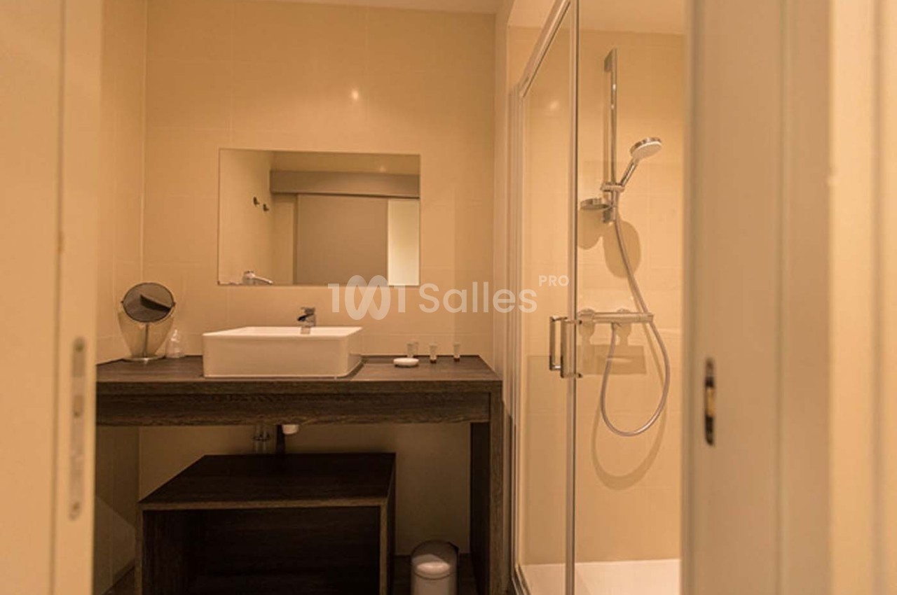 Salle de bain avec lavabo sur meuble en bois sombre, miroir rectangulaire et douche à paroi vitrée.
