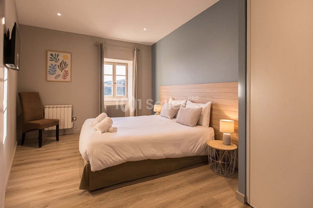 Chambre lumineuse avec lit double, table de chevet, lampes allumées, chaise et fenêtre donnant sur l'extérieur.