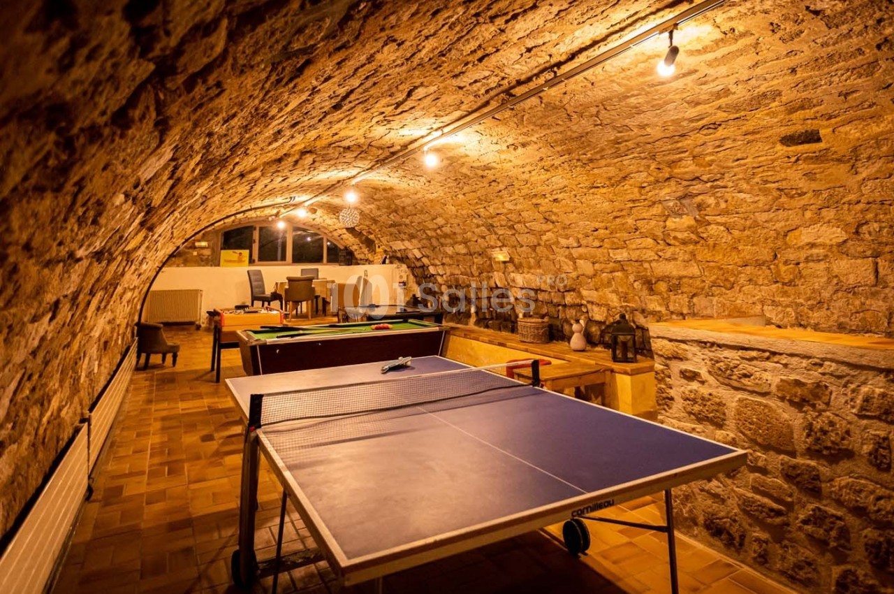 Salle voûtée en pierre avec table de ping-pong, billard et coin salon éclairés par des ampoules suspendues.