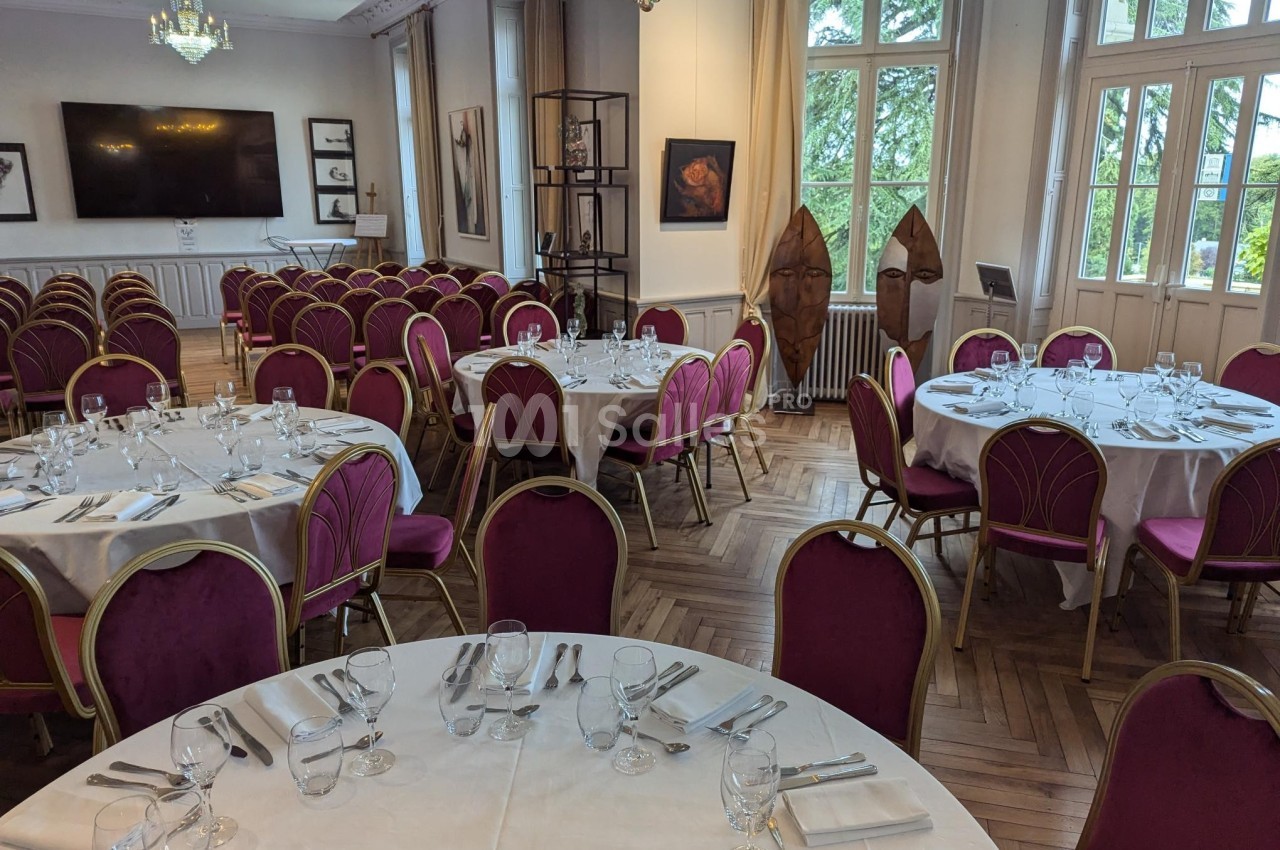 Salle élégante avec tables rondes dressées, chaises rouges, grandes fenêtres et décorations murales.