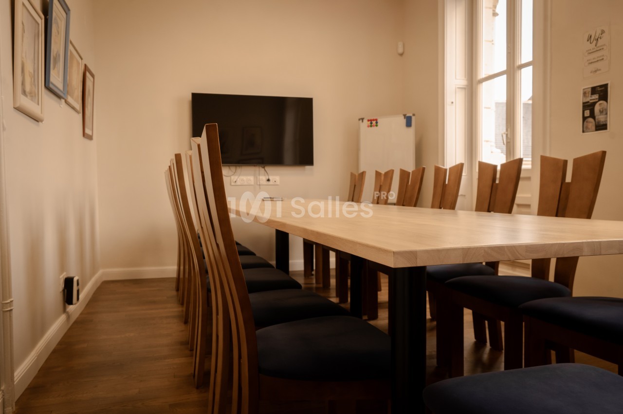 Salle de réunion avec une grande table en bois entourée de chaises, un écran mural et des fenêtres laissant entrer la…