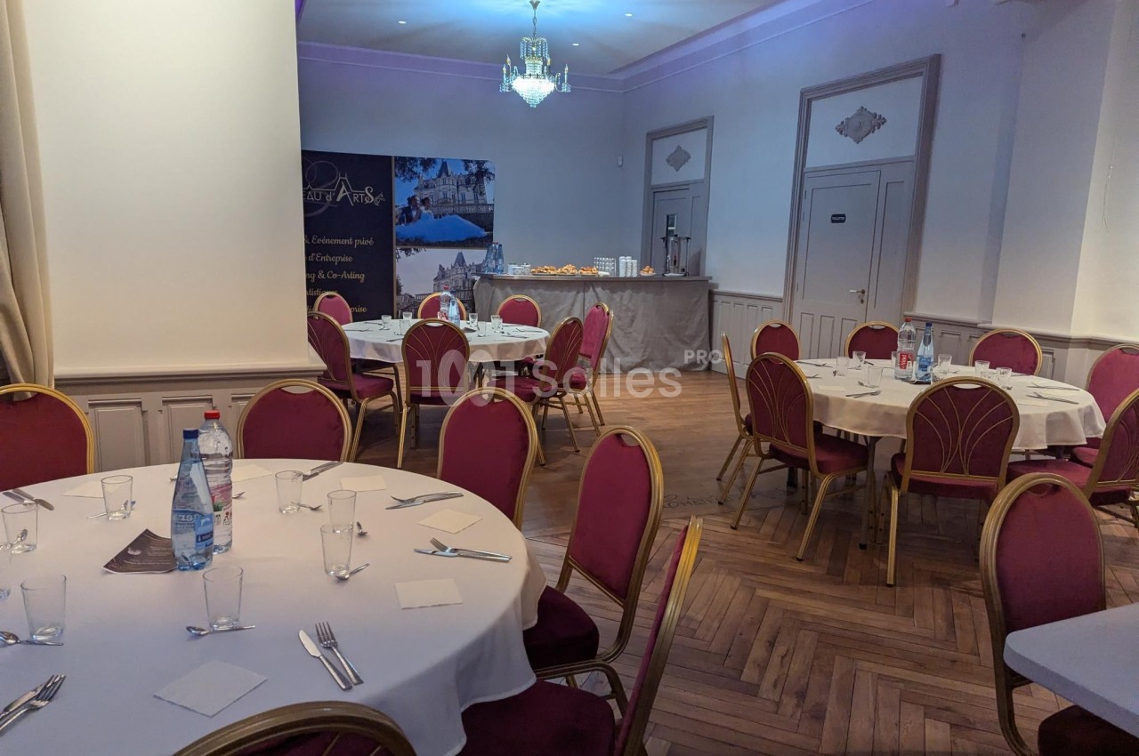 Salle de réception avec tables rondes dressées, chaises rouges et buffet en arrière-plan.