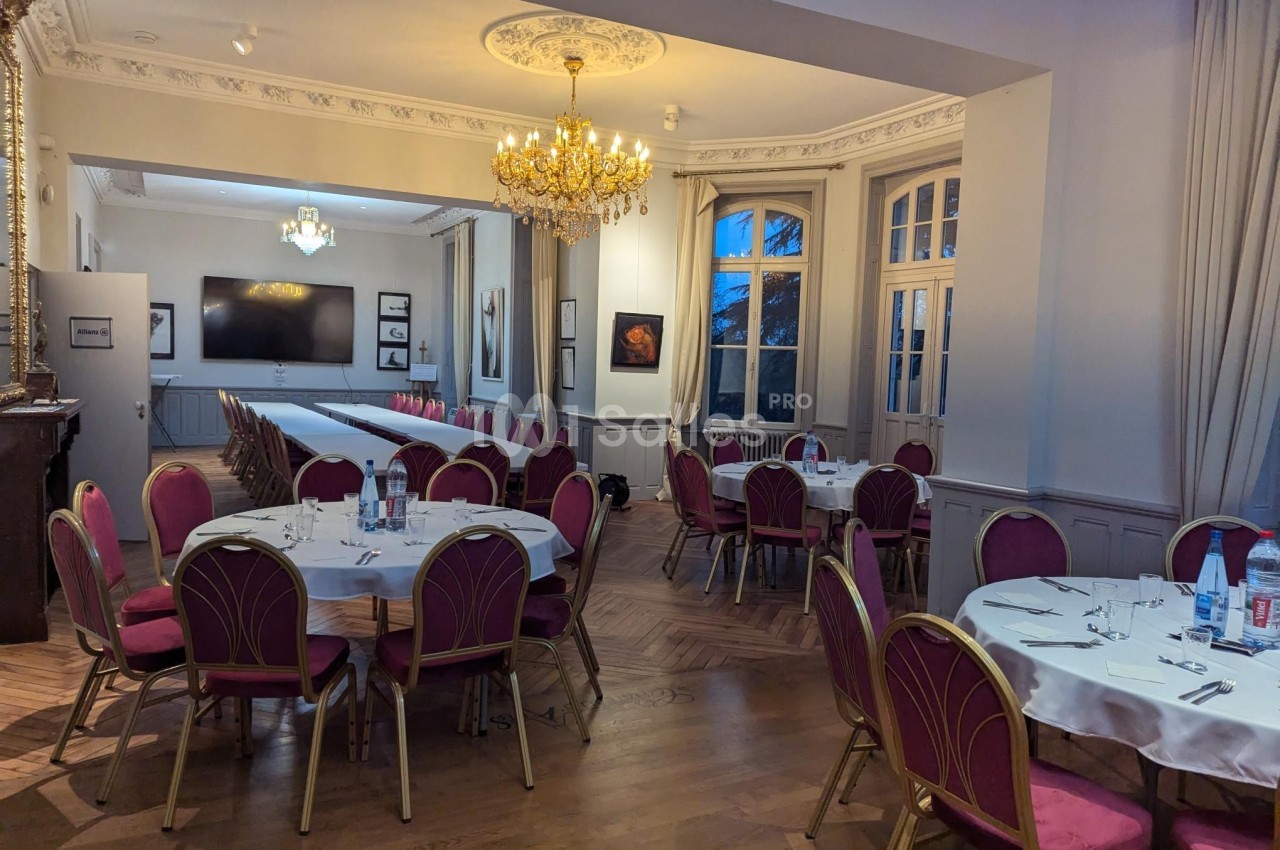 Salle élégante avec tables rondes dressées, chaises rouges, parquet en bois, lustre doré et grandes fenêtres.