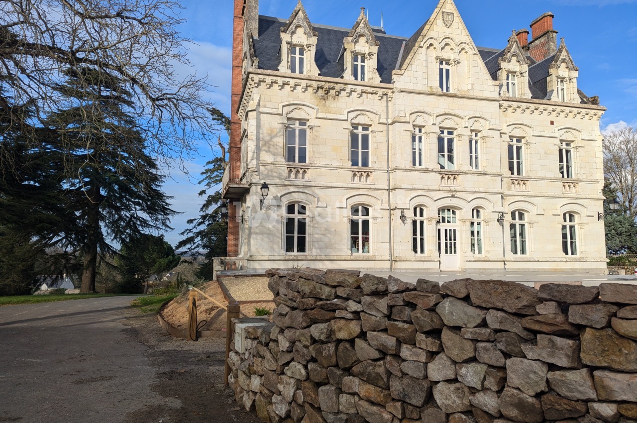 Façade d'un bâtiment en pierre de style ancien avec un mur de pierres sèches au premier plan et des arbres en arrière-plan.