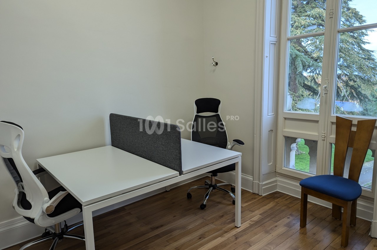 Deux bureaux blancs avec chaises, séparés par une cloison grise, près d'une fenêtre donnant sur un jardin.