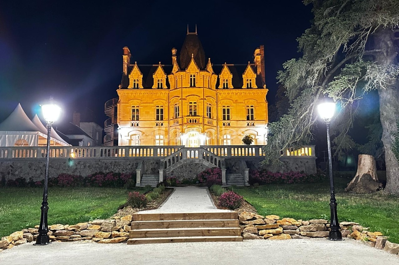 Façade illuminée d'un château de style néogothique la nuit, entourée de lampadaires et d'un jardin aménagé.