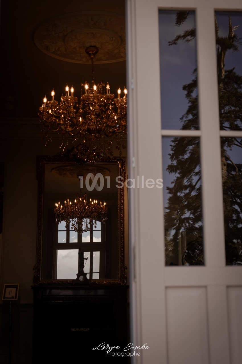Reflet d'un lustre doré et d'une fenêtre dans un miroir, avec un arbre visible à travers une porte entrouverte.