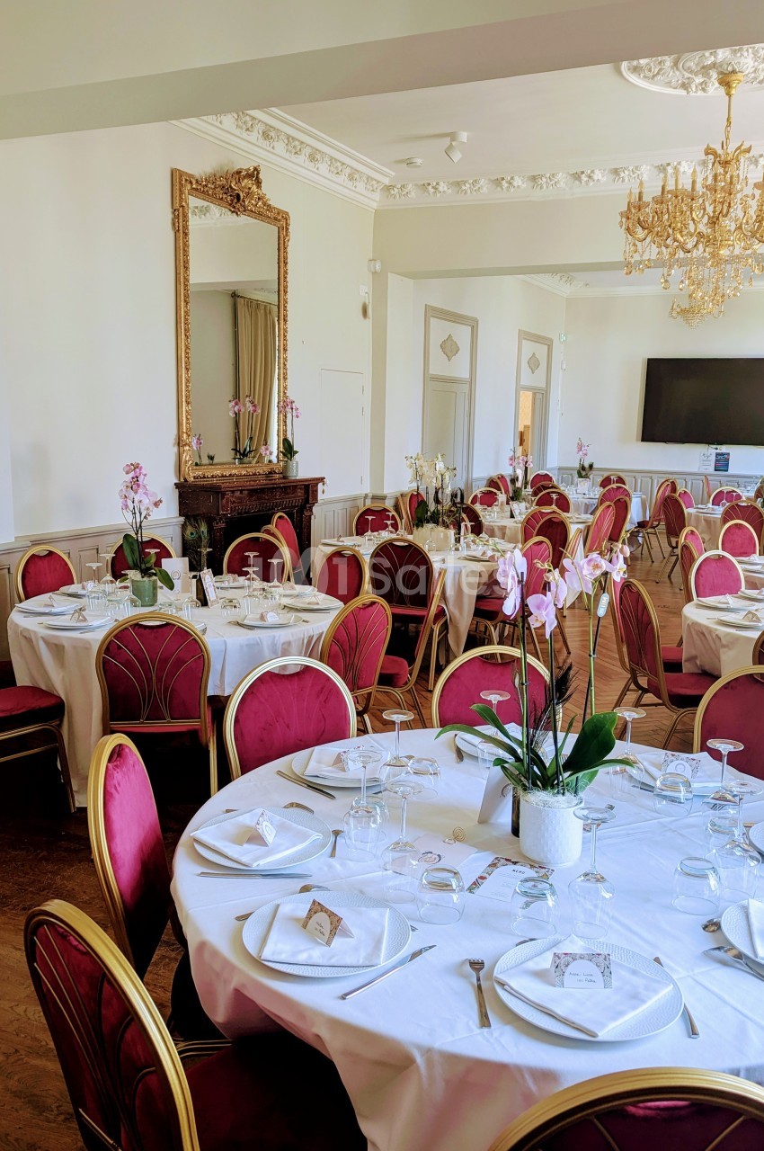 Salle de réception élégante avec tables rondes dressées, chaises rouges, miroir doré et lustre en cristal.