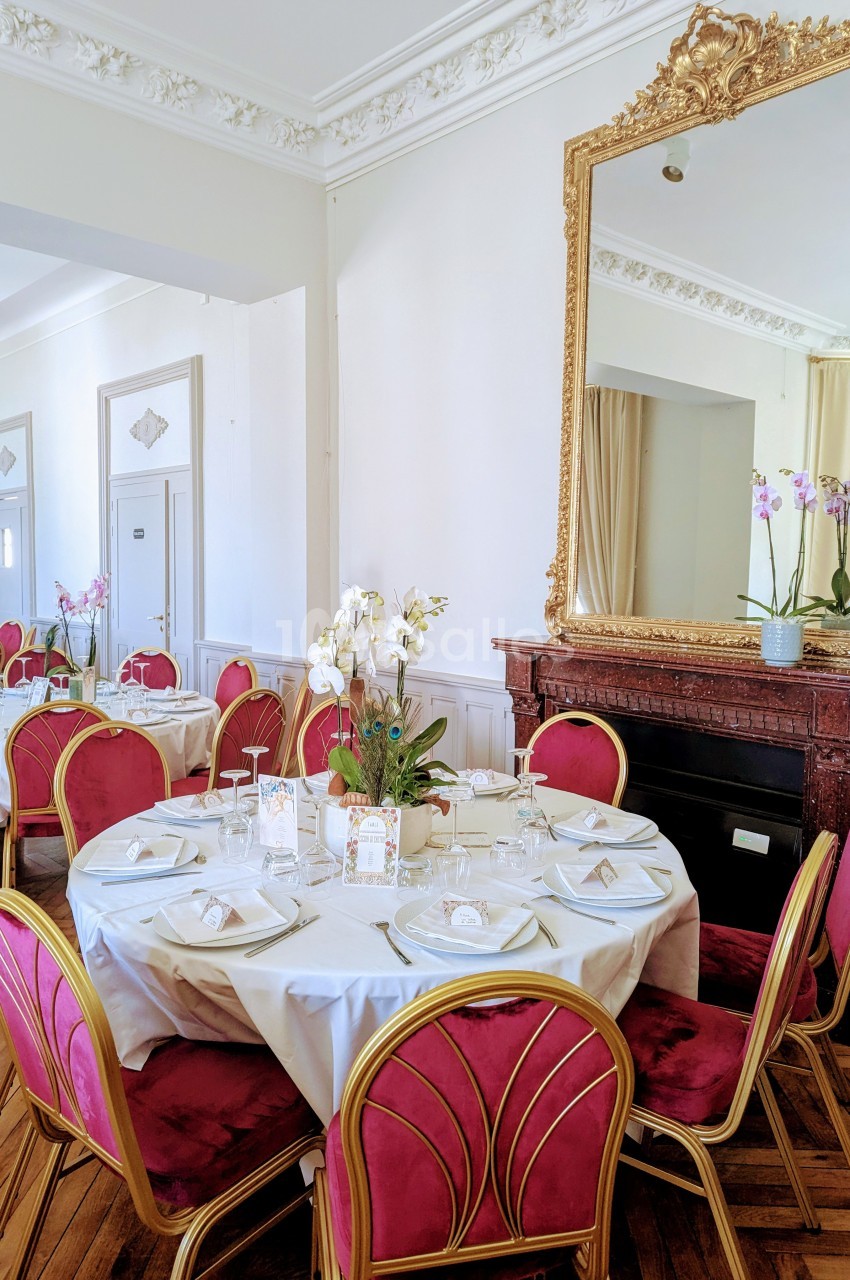 Salle élégante avec tables rondes dressées, chaises rouges et dorées, grand miroir doré et orchidées en décoration.