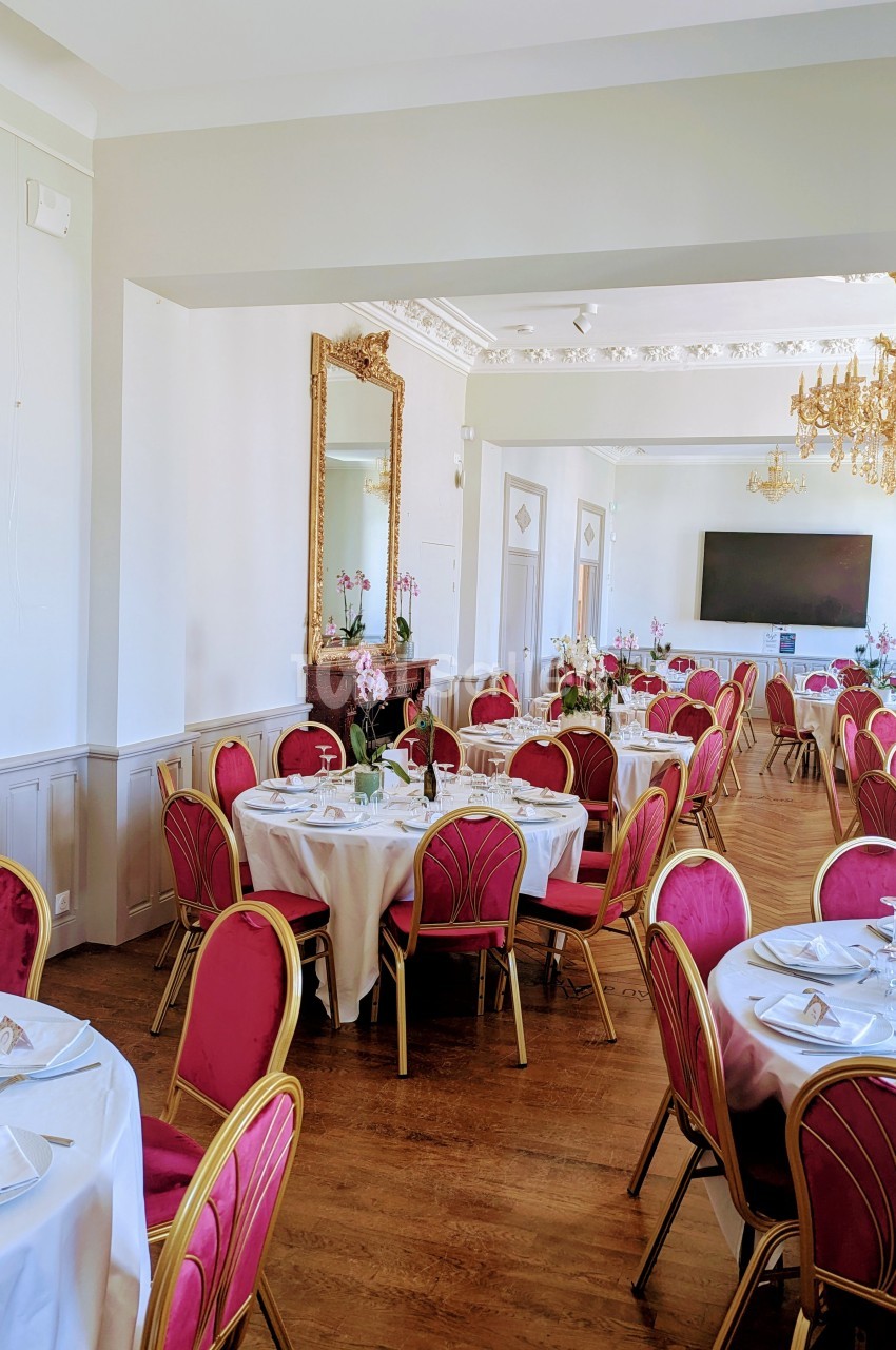 Salle de réception lumineuse avec tables rondes dressées, chaises rouges et dorées, miroir et lustre élégant.